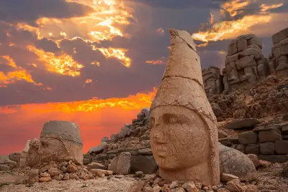 Un drammatico tramonto rosso fuoco esplode dietro una colossale testa in pietra — la divinità con corona conica e il suo volto crepato inclinato verso lo spettatore — sulla terrazza occidentale del Monte Nemrut, nuvole temporalesche che ardono in arancione e viola sopra, una seconda testa visibile a