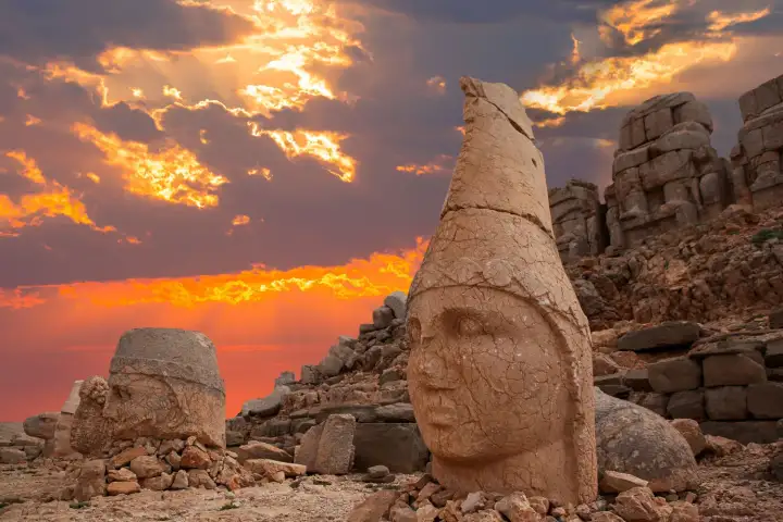 A dramatic fire-red sunset erupts behind a colossal stone head — the cone-crowned deity with its cracked face angled toward the viewer — on Mount Nemrut's west terrace, storm clouds blazing in orange and purple above, a second head visible to the left and the tumulus platform behind in Adıyaman, Tur