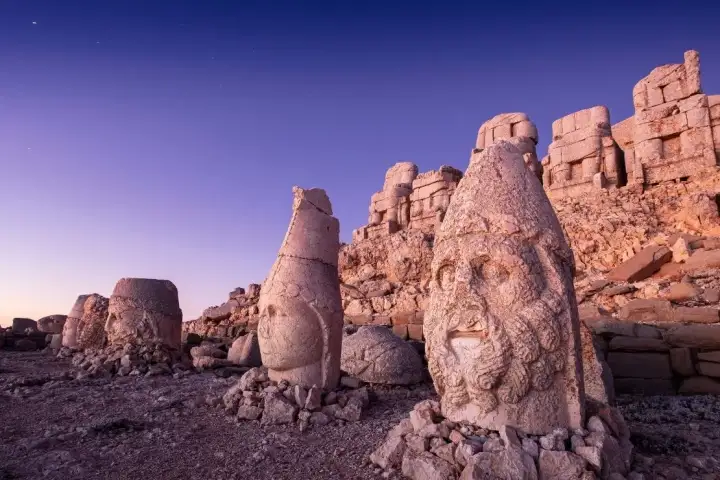 Long-exposure night photography on Mount Nemrut's west terrace shows the heavily bearded and curly-haired stone head of Heracles-Artagnes in the foreground glowing with warm raking light, while behind it a tiara-crowned deity head and additional stone figures recede toward the tumulus under a deep v