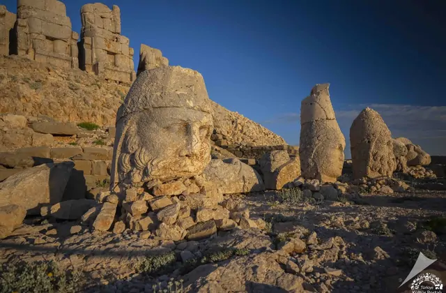 Warm side-raking early-morning light catches the deeply carved features of the Heracles-Artagnes stone head in the foreground of Mount Nemrut's east terrace, its curly beard and crown rendered in sharp relief, while two other heads and the row of intact seated deity statues stand behind it against t