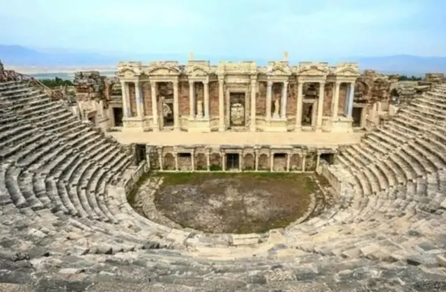The grand Roman theater of Hierapolis at Pamukkale, Turkey, seen from above with its full semicircular seating tiers and ornate scaenae frons facade rising behind the orchestra, the broad Çürüksu Valley stretching to distant mountains.