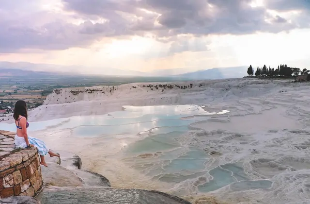 Una mujer se sienta en un saliente de piedra contemplando la vasta ladera de travertino blanco de Pamukkale al atardecer, con cipreses silueteados en la cresta y luz dorada extendiéndose sobre la llanura de Denizli.