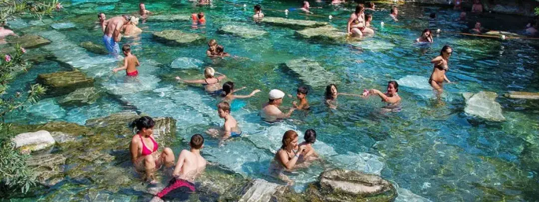 Docenas de turistas vadean y nadan entre fragmentos de columnas de mármol romano sumergidos en la Piscina Antigua de Cleopatra en Pamukkale, Turquía, con agua termal cristalina de color turquesa verde rodeada de vegetación.