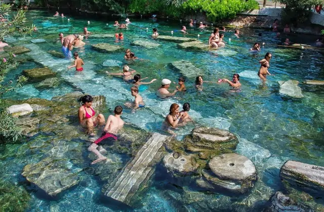 Decine di turisti guadano e nuotano tra frammenti di colonne di marmo romano sommersi nella Piscina Antica di Cleopatra a Pamukkale, Turchia, con acqua termale cristallina color verde turchese circondata da vegetazione.