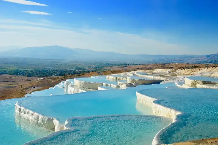 Vista panoramica dalle terrazze del Castello di Cotone di Pamukkale sulla vasta Valle di Çürüksu in Turchia, vivaci piscine termali ciano che riempiono bacini di travertino bianco smerlati in primo piano.