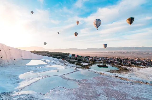Multiple colorful hot-air balloons float above Pamukkale's white travertine terraces in Turkey at sunrise, the cotton-white hillside with pale turquoise pools below the town of Pamukkale and the Denizli valley stretching to distant mountains.
