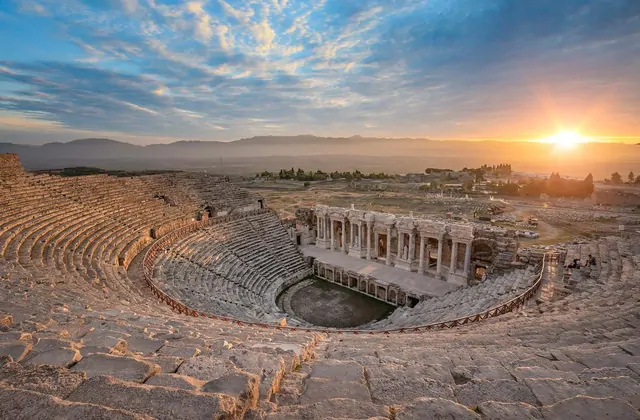 Vista aérea del teatro de Hierápolis en Pamukkale en Turquía al atardecer, las filas de asientos de piedra del anfiteatro romano antiguo dramáticamente iluminadas en naranja mientras los rayos del sol perforan las nubes.