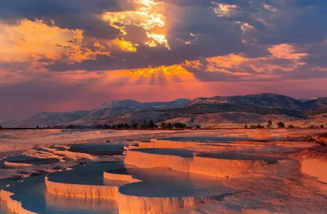 Le terrazze di travertino bianco di Pamukkale allagate di vivida acqua termale turchese sotto un drammatico cielo arancione al tramonto a Denizli, Turchia, raggi di sole che sfondano le nuvole spesse sulle montagne.