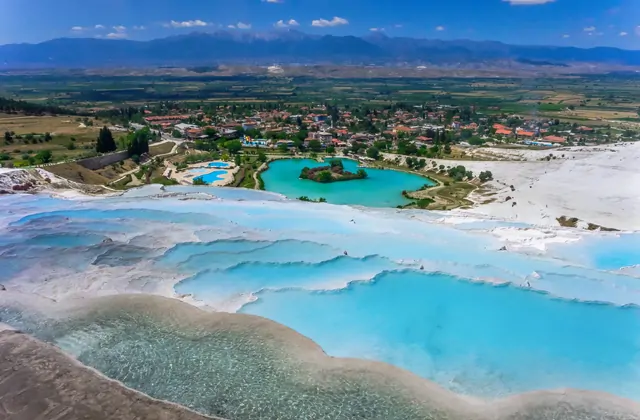 Vista aerea ampia delle terrazze di Pamukkale e del paese sotto, mostrando decine di bacini di travertino pieni di turchese a cascata lungo il fianco, con il lago termale e il villaggio visibili ai piedi e la montagna innevata dietro.