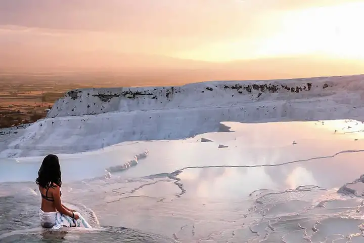 A woman with dark hair sits at the edge of Pamukkale's white travertine terraces at sunset in Turkey, facing the shallow pools that glow peach and silver under a yellow-orange sky, cypress trees and the Denizli valley far in the distance.