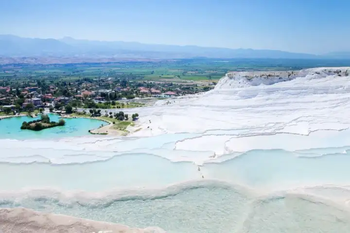 Vista aerea ampia del Castello di Cotone di Pamukkale che mostra l'intero fianco di travertino bianco con piscine poco profonde turchesi e il grande lago termale verde smeraldo sotto, con il paese di Pamukkale e la pianura di Denizli.