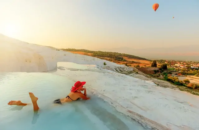 Pamukkale's white travertine terraces at dusk seen from the hillside edge, the pools cast in deep blue-violet tones as city lights glow below and a dramatic swirling sunset sky in orange and purple lights up the upper frame.