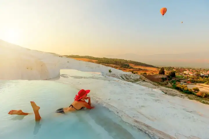 Pamukkale's white travertine terraces at dusk seen from the hillside edge, the pools cast in deep blue-violet tones as city lights glow below and a dramatic swirling sunset sky in orange and purple lights up the upper frame.