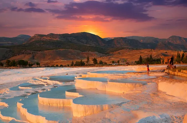 Le terrazze di travertino di Pamukkale sotto un cielo di tramonto arancione ardente in Turchia, i bordi di carbonato di calcio bianco che riflettono ambra calda mentre un gruppo di visitatori si trova al bordo lontano.