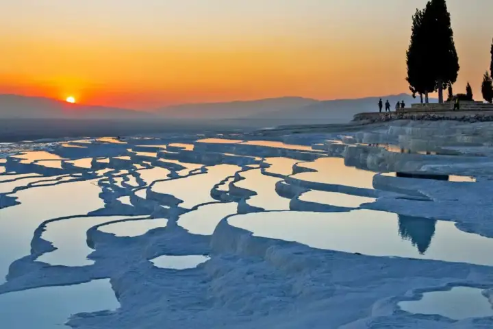 Silhouette of two tall cypress trees at the edge of Pamukkale's Cotton Castle at sunset in Turkey, the white travertine terraces catching the golden hour light as dozens of irregular pool shapes glitter below and a glowing orange horizon fades over the valley.