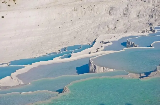 Vista aérea de las terrazas de travertino blanco de Pamukkale al anochecer mostrando acantilados irregulares de brillante agua mineral azul en cascada por niveles escalonados, la vasta ladera de piedra calcárea blanca arriba.