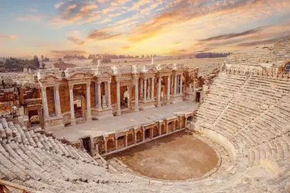The Hierapolis theater at Pamukkale, Turkey at golden hour with dramatic storm clouds breaking overhead, dramatic shafts of light illuminating the ancient stone stage facade and the curved rows of seats, the excavated ruins of Hierapolis spreading beyond.