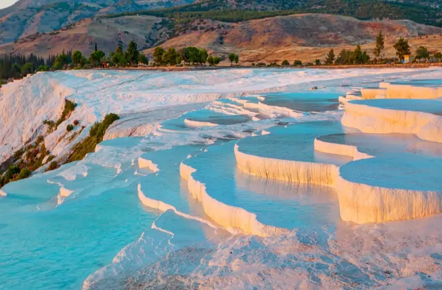 Il bordo della terrazza di travertino bianco di Pamukkale illuminato dalla calda luce dell'ora d'oro, acqua termale turchese che riempie una vasta piscina piatta in Turchia, il fianco boscoso con alberi autunnali rossi e arancioni a sinistra.