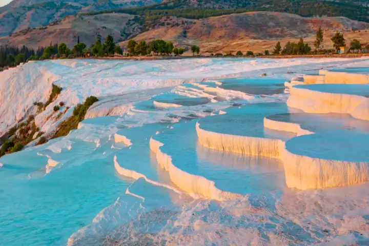 Il bordo della terrazza di travertino bianco di Pamukkale illuminato dalla calda luce dell'ora d'oro, acqua termale turchese che riempie una vasta piscina piatta in Turchia, il fianco boscoso con alberi autunnali rossi e arancioni a sinistra.