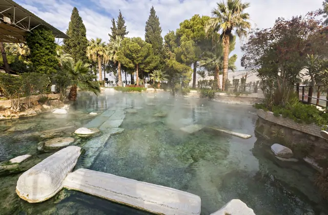 La Piscina Antica di Cleopatra a Pamukkale Hierapolis in Turchia senza nuotatori, grandi tamburi di colonne di marmo romano rotte sul fondo della limpida piscina termale fumante circondata da alte palme.