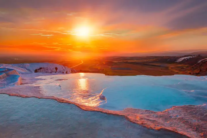 Tramonto arancione ardente sulle terrazze di travertino bianco di Pamukkale in Turchia, il sole ardente visibile sull'orizzonte mentre le piscine minerali brillano di turchese e blu.