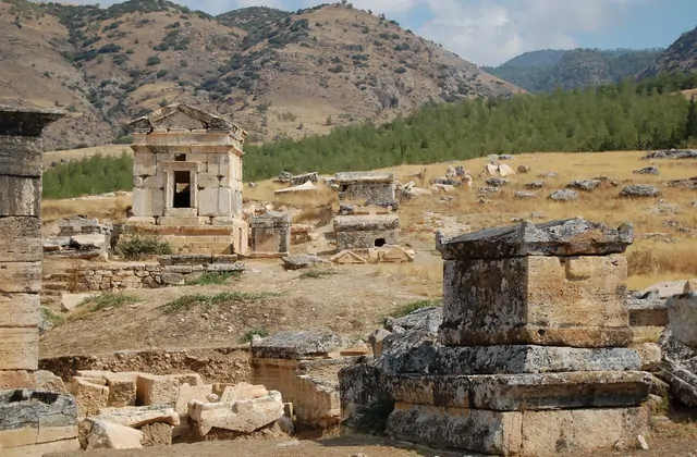 Sarcófagos de piedra y monumentos funerarios de la necrópolis de Hierápolis en Pamukkale, Turquía a distancia media, una tumba templo bien conservada con fachada de frontón en pie entre bloques de piedra dispersos sobre hierba dorada seca.