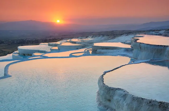 Pamukkale's white travertine terraces at a vivid orange-red sunset in Turkey, the flat sun disk hovering over the horizon and bathing the serpentine pool edges in fiery light while the Denizli Valley fades into a glowing amber haze.