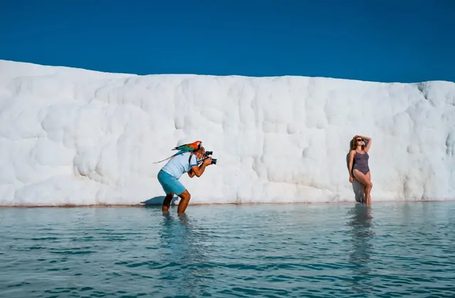 A photographer with a parrot on his shoulder photographs a woman posing in the shallow water in front of Pamukkale's brilliant snow-white travertine cliff wall in Turkey, both figures silhouetted against the towering white calcium carbonate face under deep blue sky.