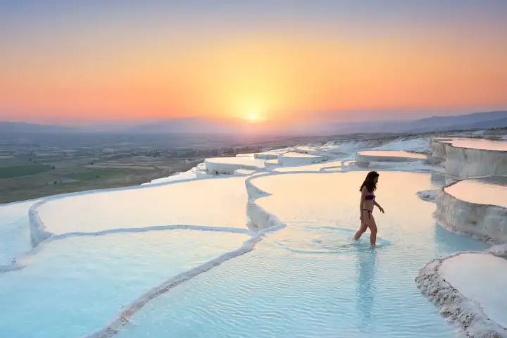A woman in a bikini walks through shallow turquoise water on Pamukkale's white travertine terraces at sunset in Turkey, the cascading calcium steps climbing behind her while the orange-pink sun drops toward the wide Denizli Valley horizon.