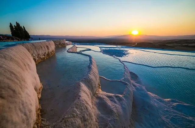 Las terrazas de Pamukkale vistas al atardecer desde el borde de la terraza con visitantes en la cresta a la izquierda lejana, dos cipreses altos silueteados contra un cielo naranja, piscinas serpenteantes turquesas brillando en primer plano.