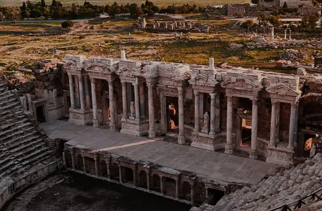 La ricca facciata scenica del teatro di Hierapolis a Pamukkale in primo piano al crepuscolo, l'arcata di colonne scolpite superiori e le nicchie ad arco con statue visibili in dettaglio, la pietra antica che brilla di ambra calda.