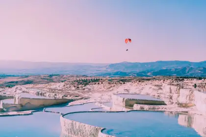 A paraglider with a vivid red canopy soars over the white travertine terraces of Pamukkale at dusk in Turkey, the pale pink and blue sky behind, flat serpentine pools below, and the Denizli mountain chain visible across the hazy valley.