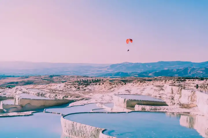 Un parapendio con una vela rossa vivace sorvola le terrazze di travertino bianco di Pamukkale al crepuscolo in Turchia, il cielo rosa pallido e blu sullo sfondo, piscine serpentine piatte sotto.