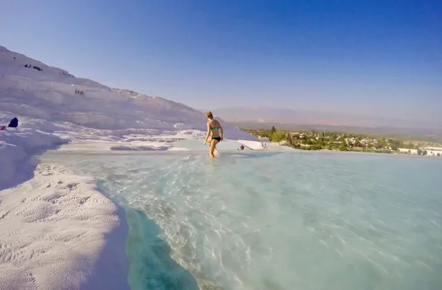 Una donna in bikini teal guada attraverso una grande piscina termica poco profonda a Pamukkale, Turchia, il fianco di travertino bianco che si innalza dietro di lei e la valle di Denizli con vegetazione visibile in lontananza.