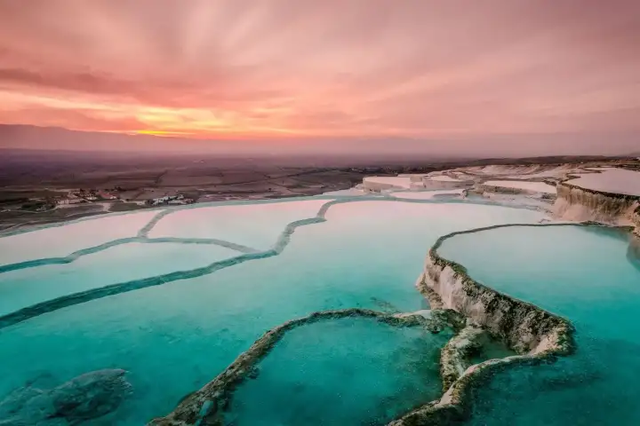 Wide aerial view of Pamukkale's pink-tinged travertine terraces at pink sunset in Turkey, vivid teal-green thermal pools filling large irregular basins divided by white calcium ledges, the harvested Denizli plain visible under a pink and red sky.