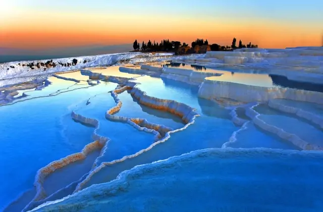 Panoramic sunset view of Pamukkale's terraces in Turkey with a deep blue glowing thermal pool in the foreground, the white calcium terrace edges catching orange sunset light as cypress trees cluster on the upper ridge and Denizli glows in the distance.