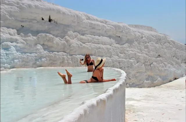 Two women in swimwear relax in a shallow natural pool at the edge of Pamukkale's white travertine terraces in Turkey, one sitting and photographing, the other reclining against the curved calcium rim as the bright midday sun bleaches the formations.