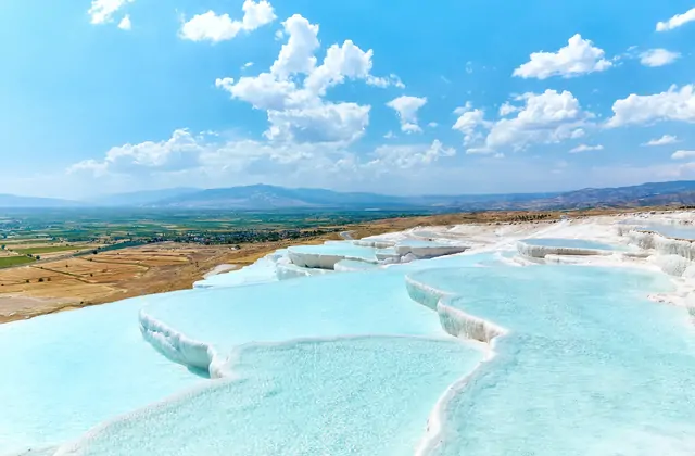 Las amplias terrazas blancas de Pamukkale con brillantes piscinas turquesas bajo un profundo cielo azul con cúmulos blancos en Turquía, la vista de la ladera desde arriba mostrando la extensión completa de las formaciones de calcio escalonadas.