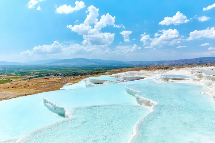 Las amplias terrazas blancas de Pamukkale con brillantes piscinas turquesas bajo un profundo cielo azul con cúmulos blancos en Turquía, la vista de la ladera desde arriba mostrando la extensión completa de las formaciones de calcio escalonadas.