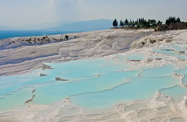 Le terrazze di Pamukkale al crepuscolo con le luci del paese che iniziano ad illuminarsi, una grande cascata di travertino bianco in primo piano nella distanza media mentre piscine blu cobalto poco profonde si estendono in primo piano.