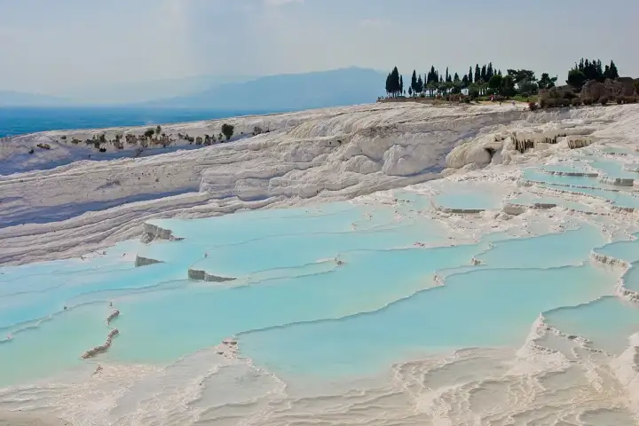 Pamukkale's terraces at dusk with the town lights beginning to illuminate, a large white travertine cascade face rising in the middle distance while shallow cobalt blue pools stretch in the foreground and a fiery orange and violet sky illuminates the horizon.