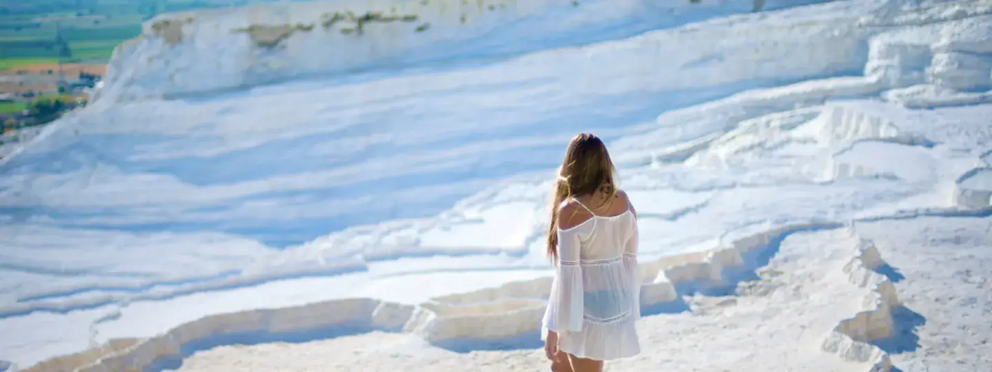 Una mujer con un vestido blanco fluido está descalza al borde de una piscina termal de Pamukkale en Turquía, mirando la ladera de travertino blanco escalonada y el amplio Valle de Denizli verde muy abajo.
