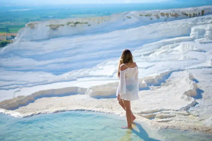 A woman in a flowing white dress stands barefoot at the edge of a Pamukkale thermal pool in Turkey, looking over the tiered white travertine hillside and the broad green Denizli Valley far below under a bright midday sky.