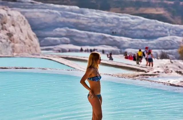 Una donna in bikini blu si trova sul bordo di una grande piscina di travertino turchese a Pamukkale in Turchia, altri visitatori e piscine visibili dietro di lei in lontananza mentre il fianco di calcio bianco si innalza.