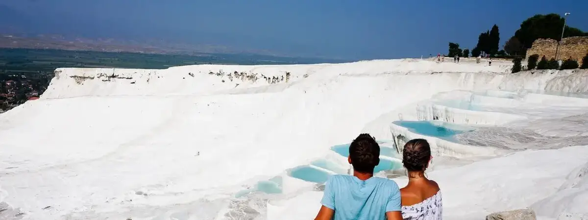 Una coppia si abbraccia nelle acque poco profonde della piscina termica di Pamukkale in Turchia, la maestosa parete di travertino bianco che si innalza dietro di loro mentre gli abeti costeggiano il bordo superiore.