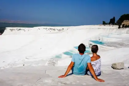 A couple embraces in the shallow thermal pool water at Pamukkale in Turkey, the towering white travertine cliff face rising behind them while fir trees line the upper edge and scattered visitors can be seen on the white terraces further back.