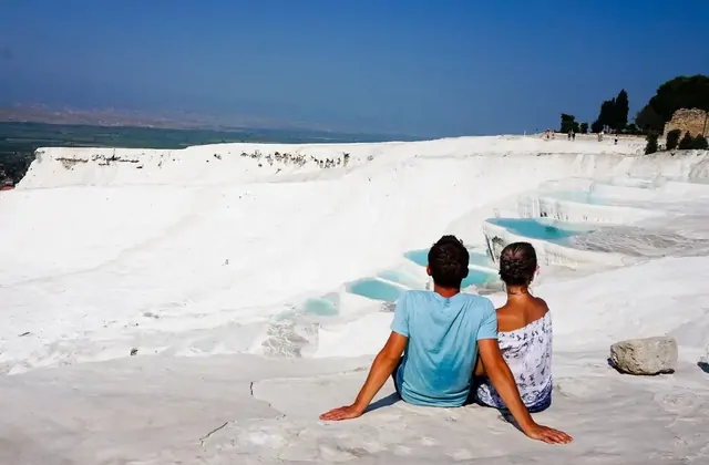 A couple embraces in the shallow thermal pool water at Pamukkale in Turkey, the towering white travertine cliff face rising behind them while fir trees line the upper edge and scattered visitors can be seen on the white terraces further back.