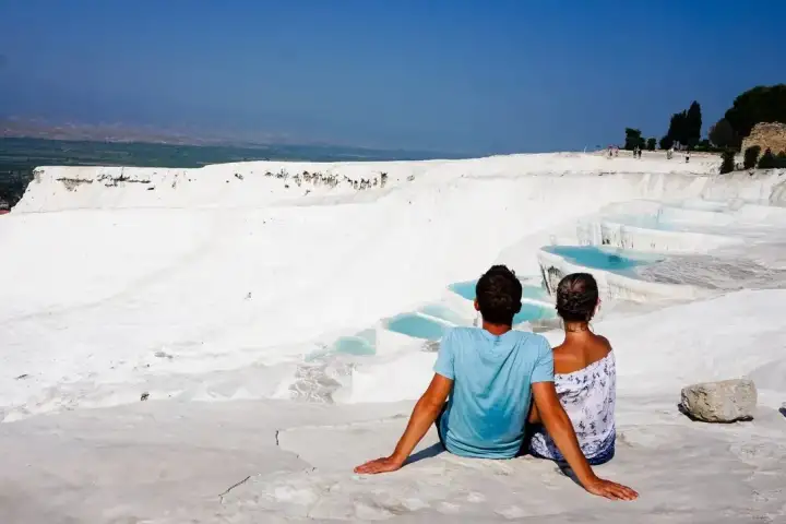 A couple embraces in the shallow thermal pool water at Pamukkale in Turkey, the towering white travertine cliff face rising behind them while fir trees line the upper edge and scattered visitors can be seen on the white terraces further back.