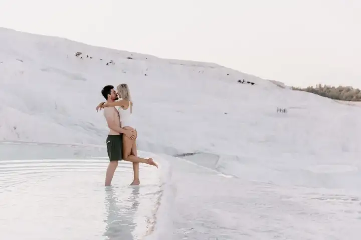 A couple shares a romantic moment in the clear thermal pool water at the base of Pamukkale's white travertine cliff in Turkey, the man holding the woman while she laughs, the vast white hillside and distant tree line framing them in the background.