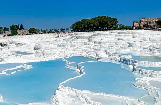 Pamukkale's stepped travertine terraces in Turkey under a bright blue sky, wide turquoise thermal pools separated by flat white calcium ridges descending in orderly tiers, tourists visible walking the upper terrace as pine trees and ancient Hierapolis ruins appear at the top.